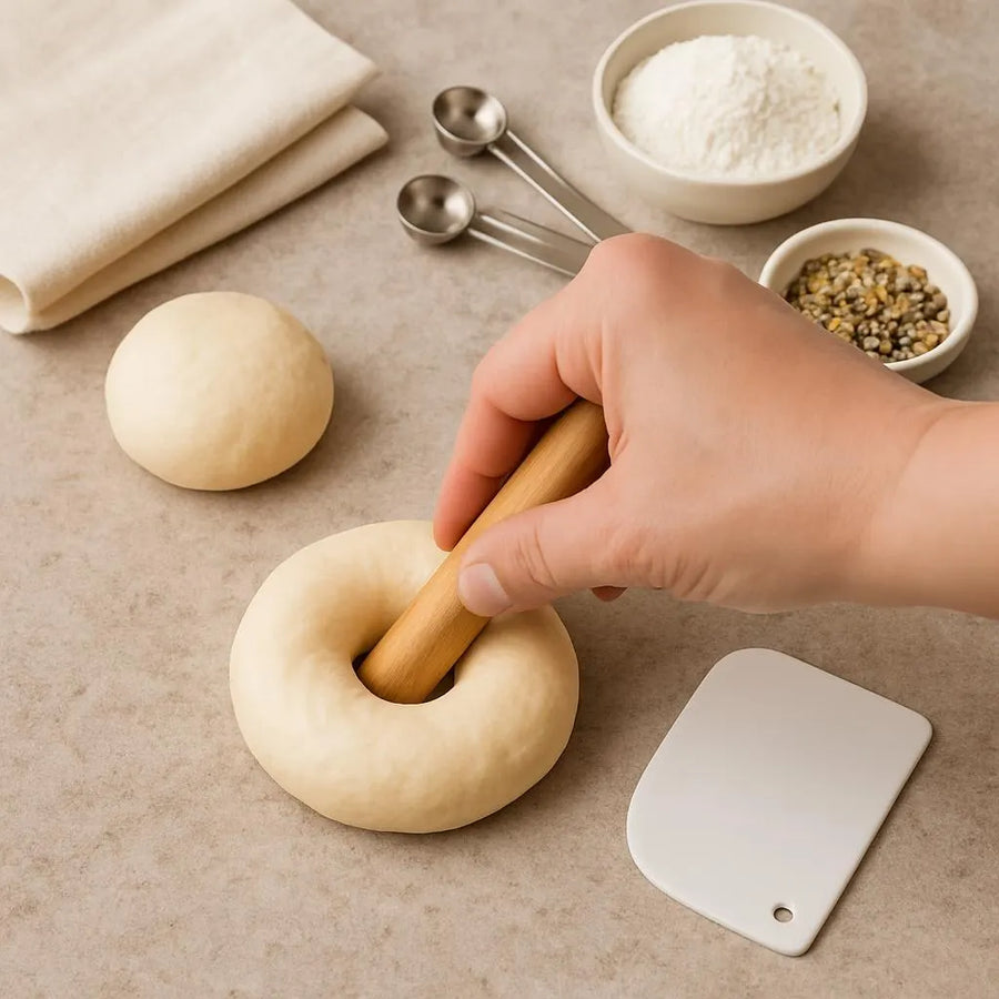 Hand using a wooden bagel-shaper dowel to punch a hole through a portioned piece of bagel dough.