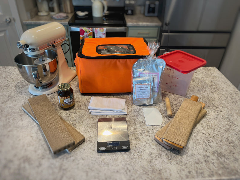Bagel-making gifts laid out on a counter: making kit, cedar board, proofing box, container, scale, stand mixer, malt syrup, and baking steel.