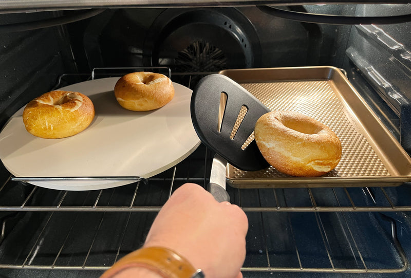 Spatula rescuing a bagel from a hot pizza stone onto a room-temperature sheet pan mid-bake.