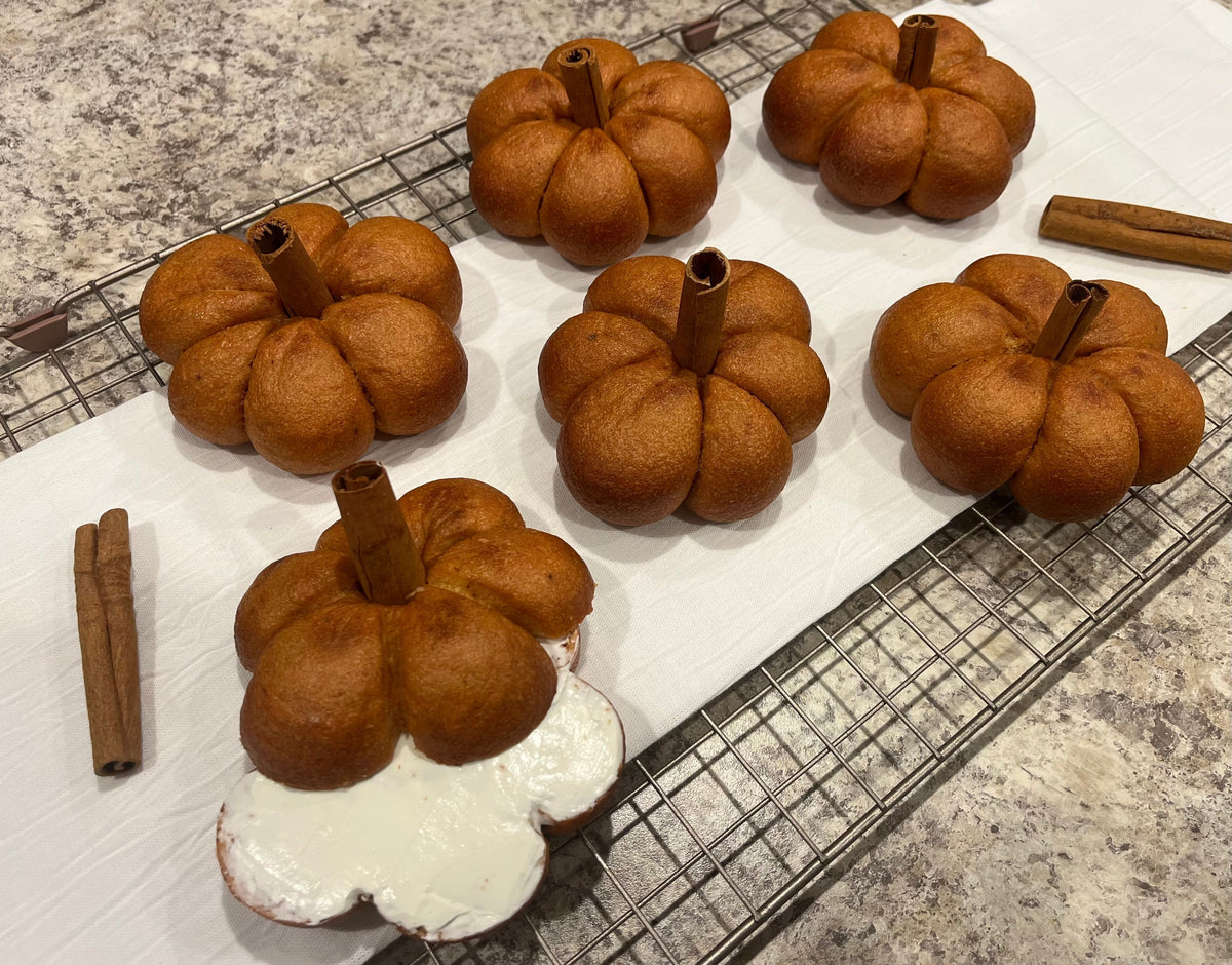 Six pumpkin-shaped bagels with cinnamon stick stems on a wire rack, one spread with cream cheese.