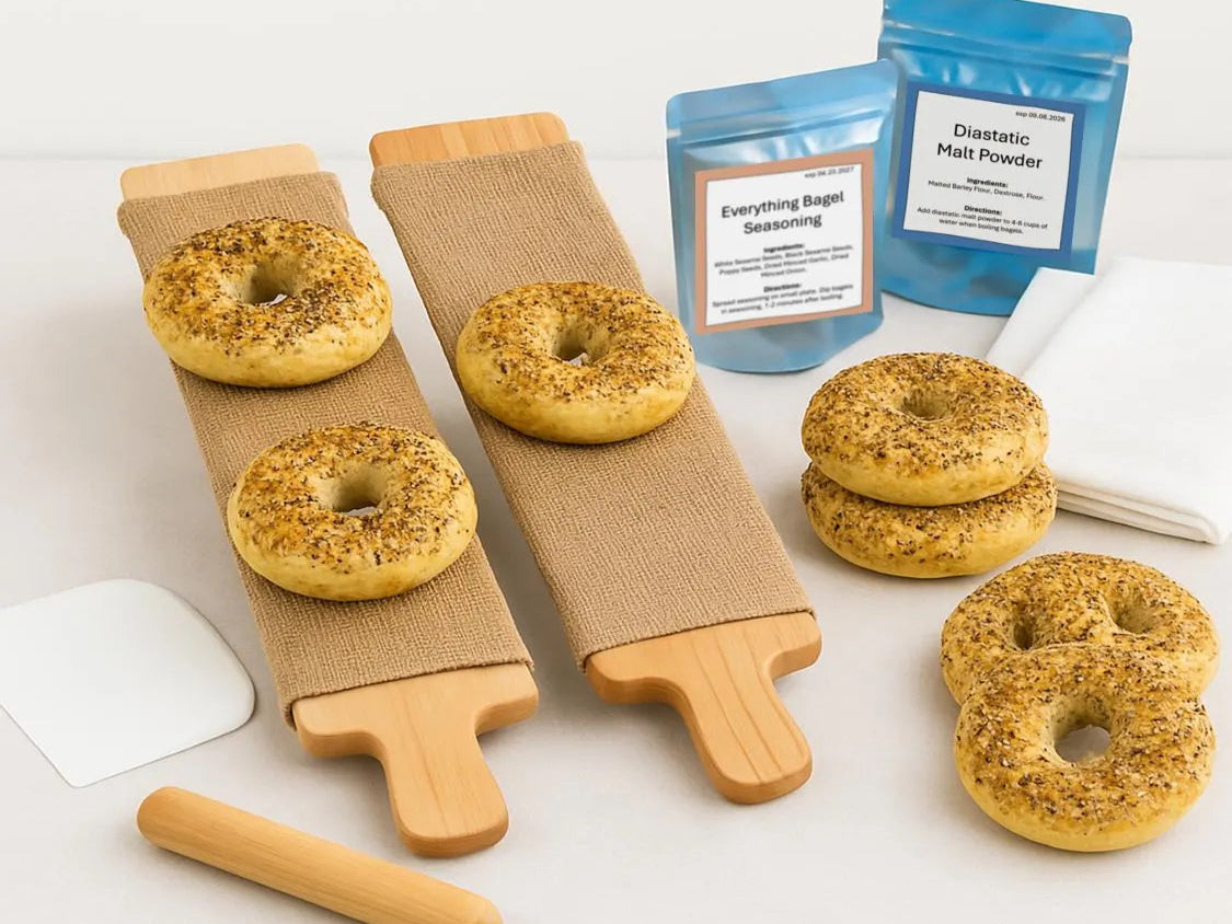 Two cedar bagel boards laid out next to a bagel-making kit's tools, ingredient bags, and recipe card on a clean surface.